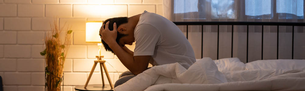 Younger man with his head in his hands on the side of his bed, suffering from insomnia